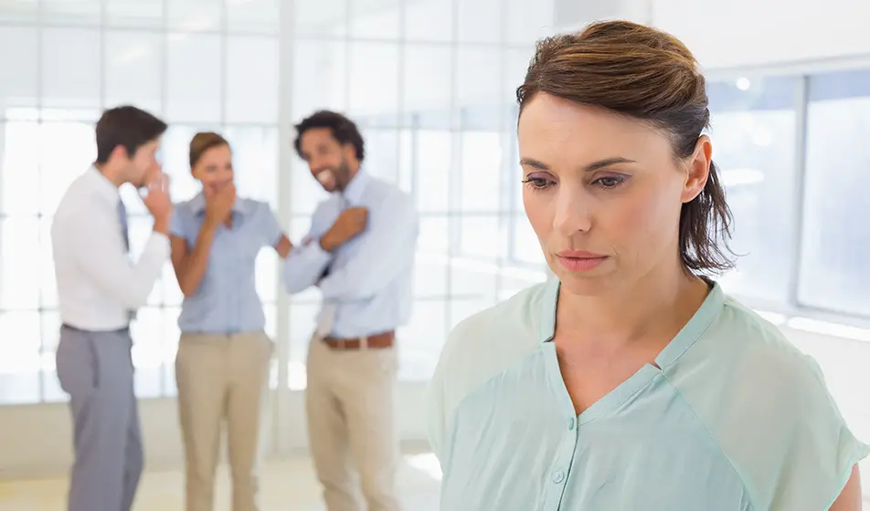 A woman stands apart from a group looking forlorn, whilst a group of colleagues appear to be talking and laughing, set apart in the background.