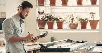 A man is writing notes on a clipboard whilst standing in front of a paper guillotine. He is near a window which has two shelves on it full of terracotta plant pots.