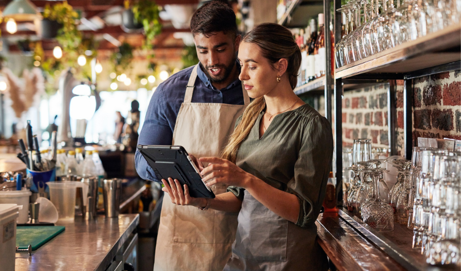 Two hospitality staff members review a tablet at a bar while managing staggered shift patterns.