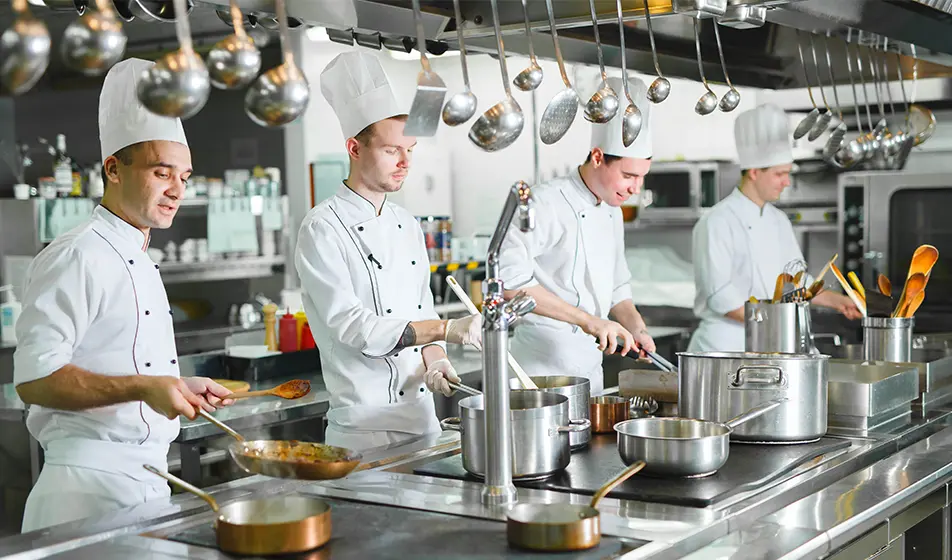 Four chefs are cooking next to each other in a restaurant kitchen. They are surrounded by silver pots  and pans. There are lots of silver ladles hanging from the ceiling.
