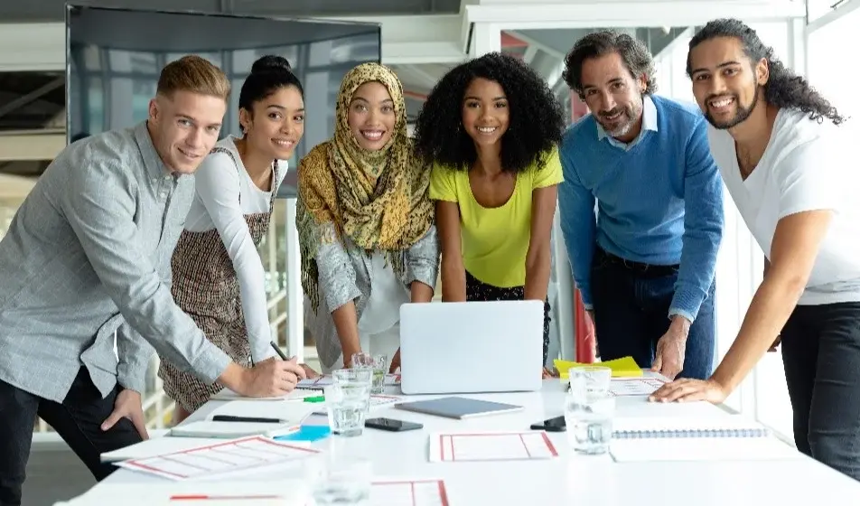 Six colleagues stood around a table together. They have a laptop in front of them which is surrounded by lots of paperwork and glasses. They are all smiling at the camera.