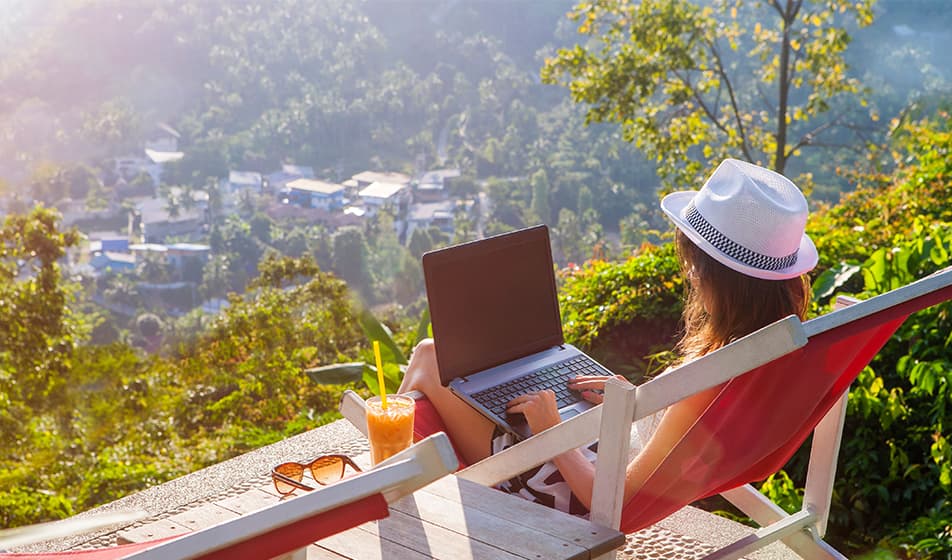A woman is sat on a red deck chair on a wooden balcony which overlooks a forest and small village. She is wearing a white hat and has a laptop on her lap. Next to her is an orange drink with a yellow straw in it. The sun is shining down on her.