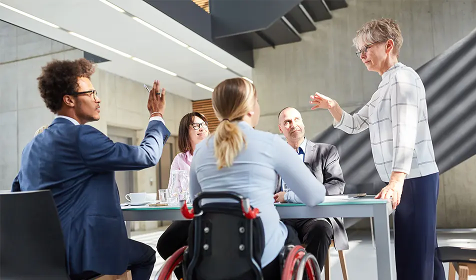 Four people are sat at a glass table. One person is in a wheelchair. One person is standing at the top of the table and is talking to the others.