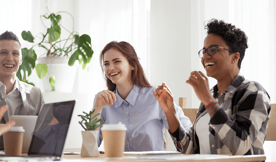 Three colleagues are sat at a table together, having a conversation and laughing. The table has takeaway coffee cups and a laptop on it. Behind one of the colleagues is a Swiss Cheese plant.