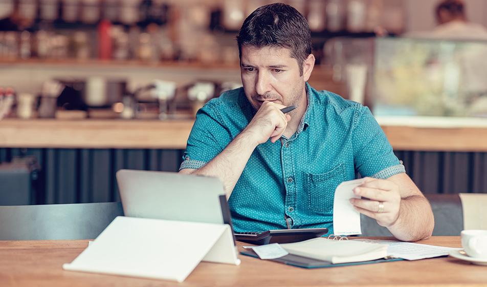 A man in a blue shirt is sat at a table in a coffee shop. His laptop is in front of him, as well as a folder full of paperwork and receipts. He is managing his expenses. 