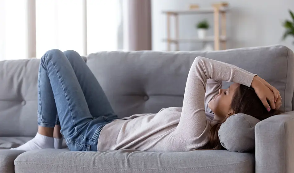 A woman is suffering from fatigue. She is lying down on a grey sofa with her hands on her head and her eyes closed.