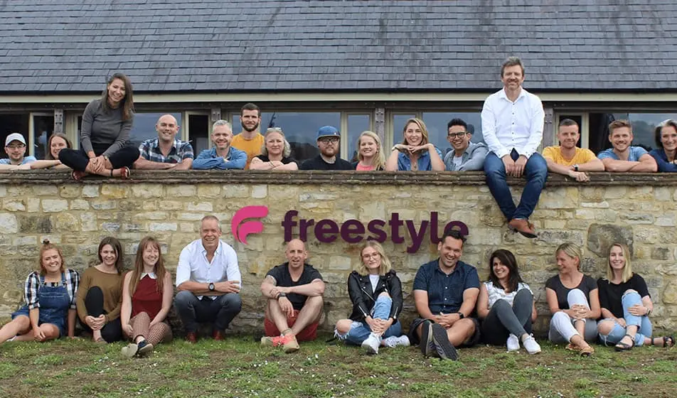 The team of Freestyle all sat together outside their office. Some of them are sat on a brick wall and some of them are sat against it.