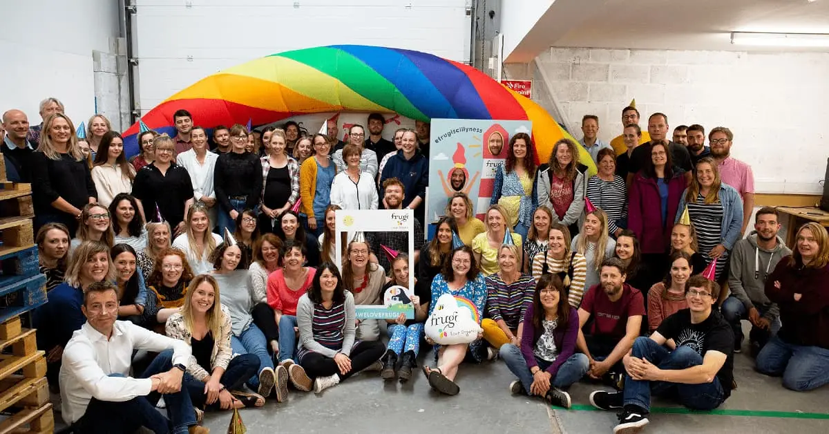 The team of Frugi all sat together in their office. There is an inflated rainbow behind the team. Everyone is smiling at the camera.
