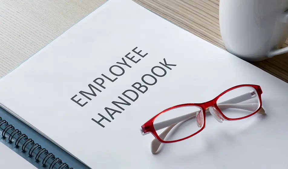 A white book is placed on a wooden table next to a coffee cup. Written on the front cover are the words "Employee Handbook" in black capital letters. On top of the notebook are some red glasses.