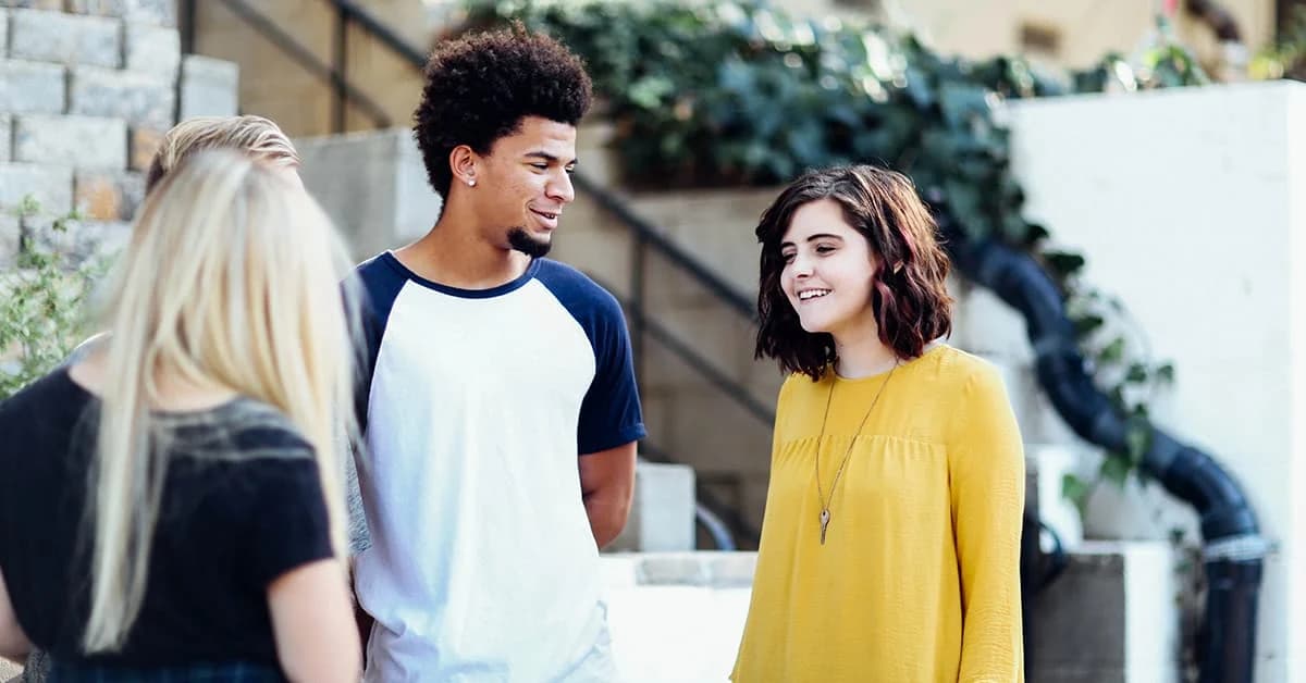 Three people are stood talking outside a building. Two of them are women, one is a man. One of the women has brown curly hair and is wearing a bright yellow jumper. The other is looking away from the camera and wearing all black. The man has is wearing a blue and white t-shirt and smiling at the woman in yellow.