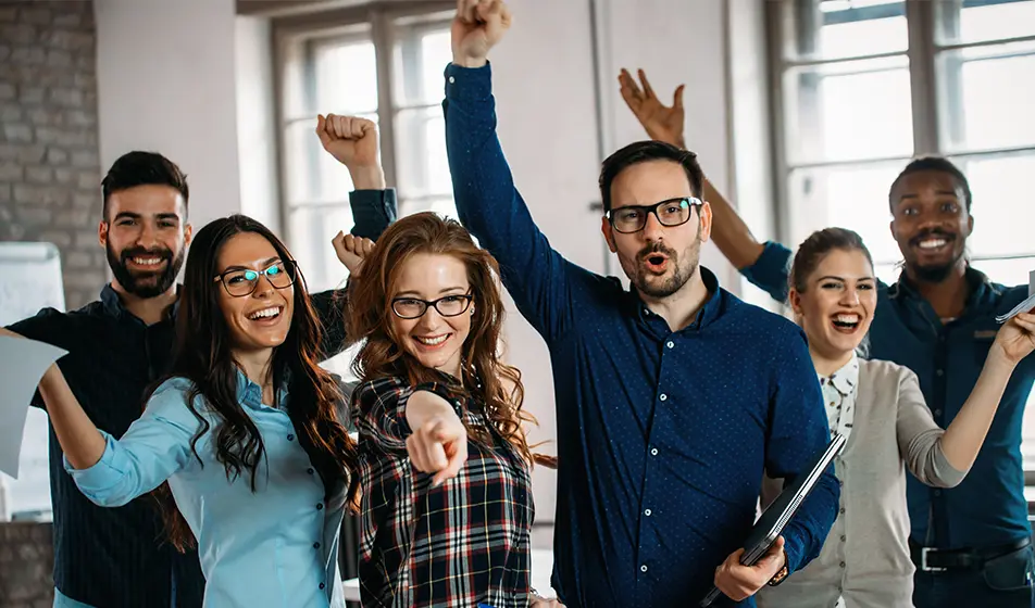 Six happy colleagues are cheering and pointing at the camera. They are celebrating.