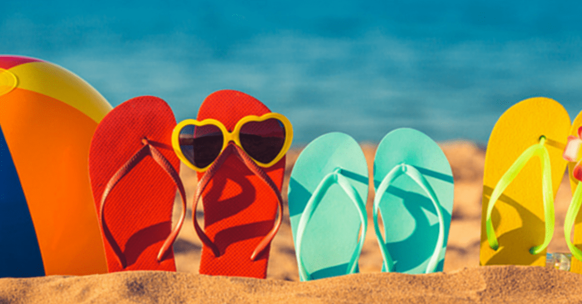 Three pairs of flip flops are standing up in the stand. The first pair of flip flops are bright red and has yellow heart-shaped sunglasses balancing on them. The second pair of flip flops are a bright light blue colour. The last pair of flip flops are bright yellow. Next to them is a colourful beach ball. In the background, there is the sea.