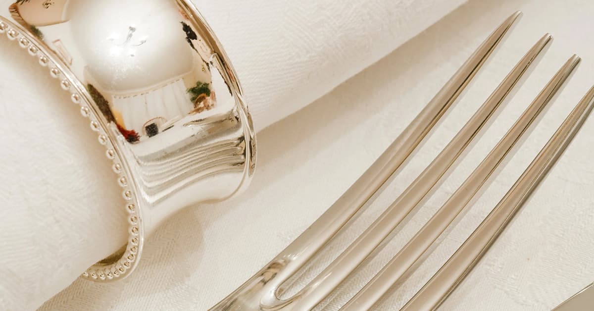 A zoomed in image of a silver fork next to a white napkin which has a thick silver ring around it.