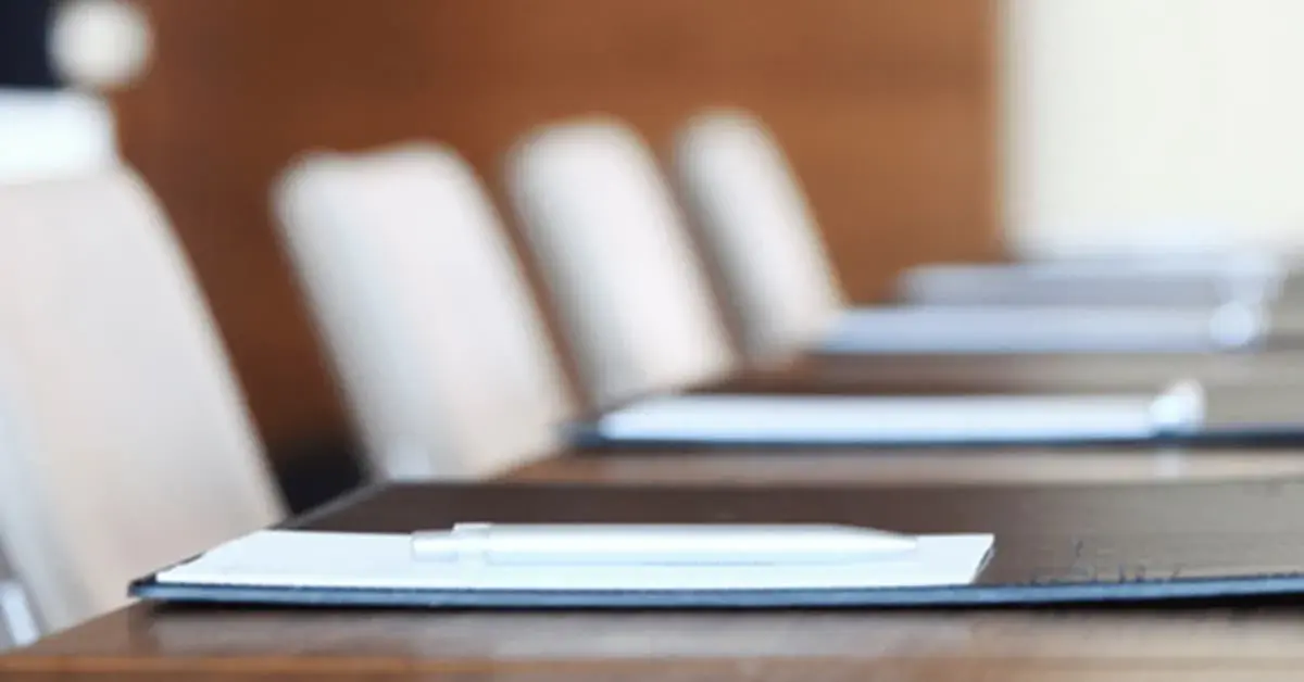 An image of a table with empty cream cushioned chairs around it. The table has black placemats on it with paperwork and pens.