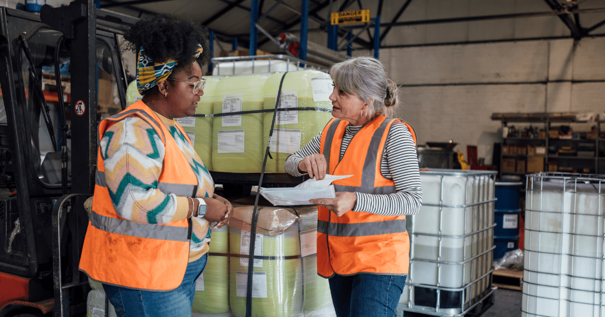 Two employees in high-visibility vests review paperwork together in a warehouse, focusing on workplace health and safety.