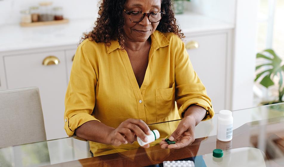 A woman sits in her kitchen, tipping some pills from a medication bottle into her hand. There's a blister pack of tablets near her, as well as another bottle of medicine. She appears to be suffering from a long-term health condition.
