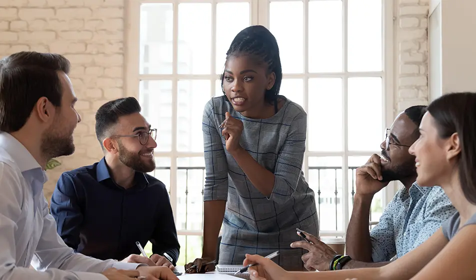 Five colleagues are working together around a table. One of the colleagues is stood up and the rest of them are sat down. All the colleagues sat down are smiling and listening to the one stood up.