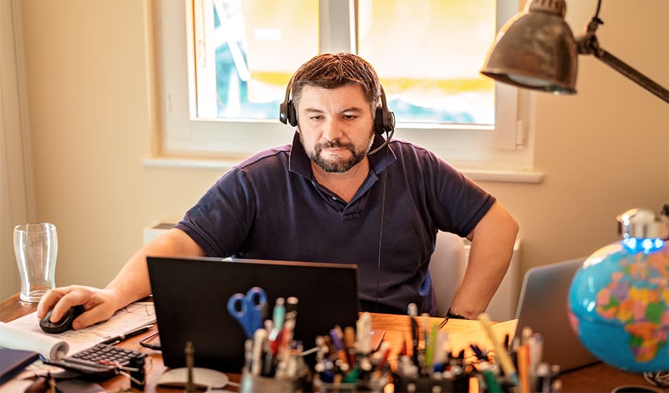 A man is working from home during Lockdown. He is sat at his desk with headphones on and his computer in front of him. On his desk there is also a globe of the world, lots of pencil pots, a calculator and an empty glass.