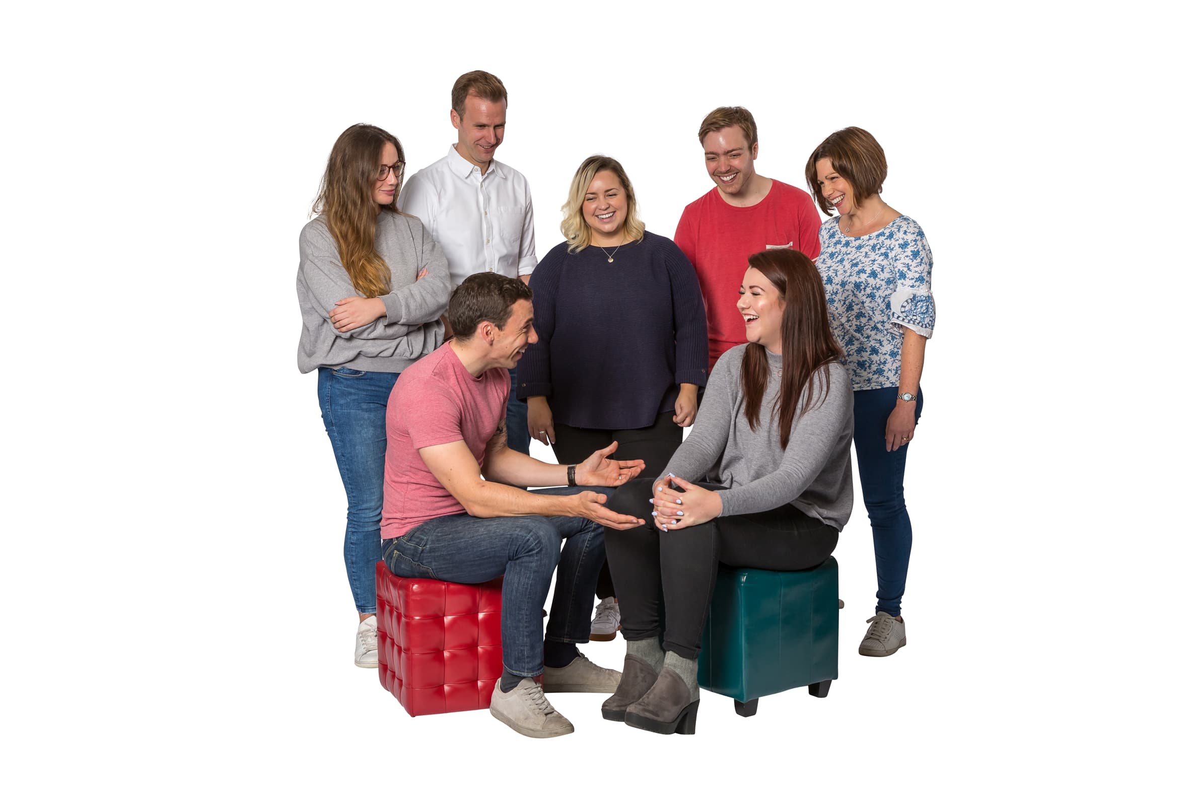A team of seven people laughing together. Two of the colleagues are sat on cushioned stools whilst the rest of the team surround them.