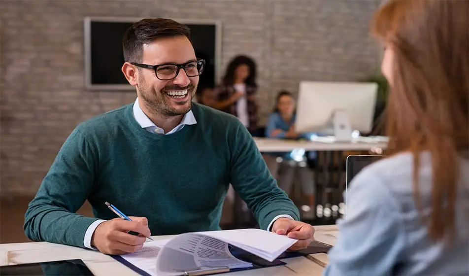 A man  and a woman are sat at a desk having an interview. The man who is hosting the interview is wearing a green jumper and is smiling at the woman across from him whilst he looks through her documents.