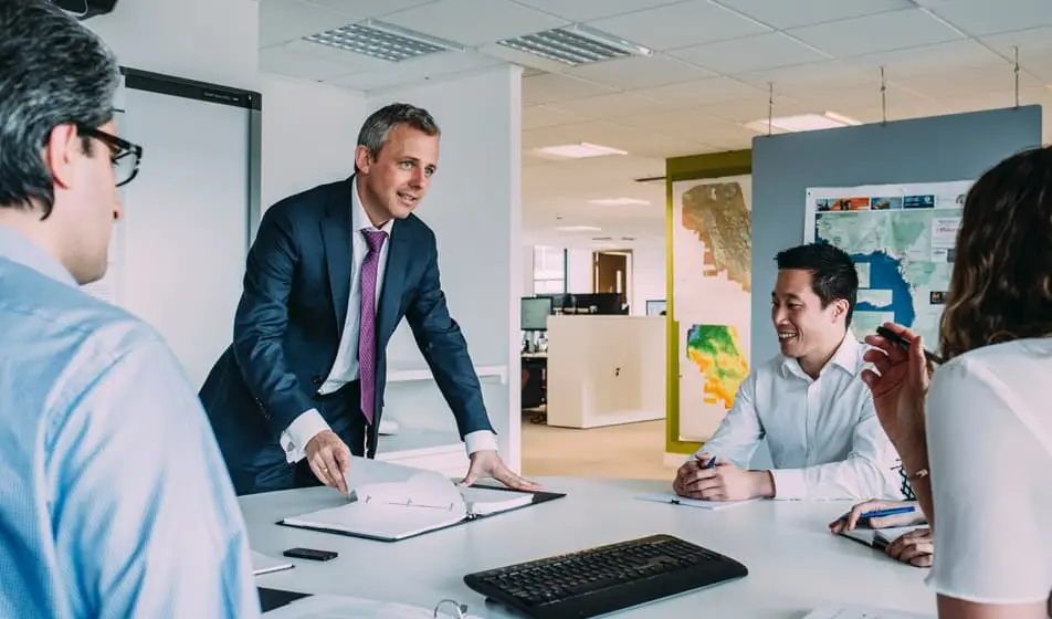 A manager is hosting a meeting with his employees who are sitting around a white table. On the table there is a keyboard and lots of notebooks.