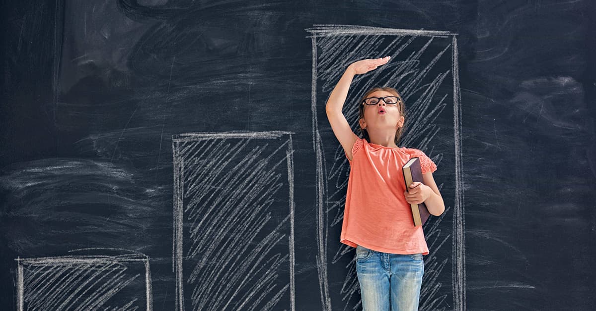 A girl is standing against a chalkboard. On the chalkboard there is a giant bar chart which includes 3 different bars. These bars are organised in height order and the girl is standing against the tallest bar which is a little taller than her.