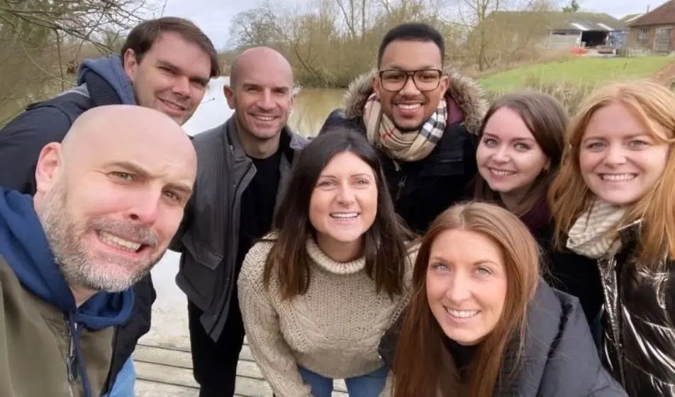 A team of eight people enjoying a walk outdoors. They are stood on a wooden path which stands over a river. They are all wrapped up in jumpers and scarves.