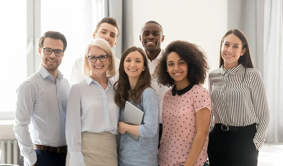 Seven colleagues are stood in an office with the natural light shining through the windows onto them.  They are all looking and smiling at the camera. The team is made up of people from different generations.