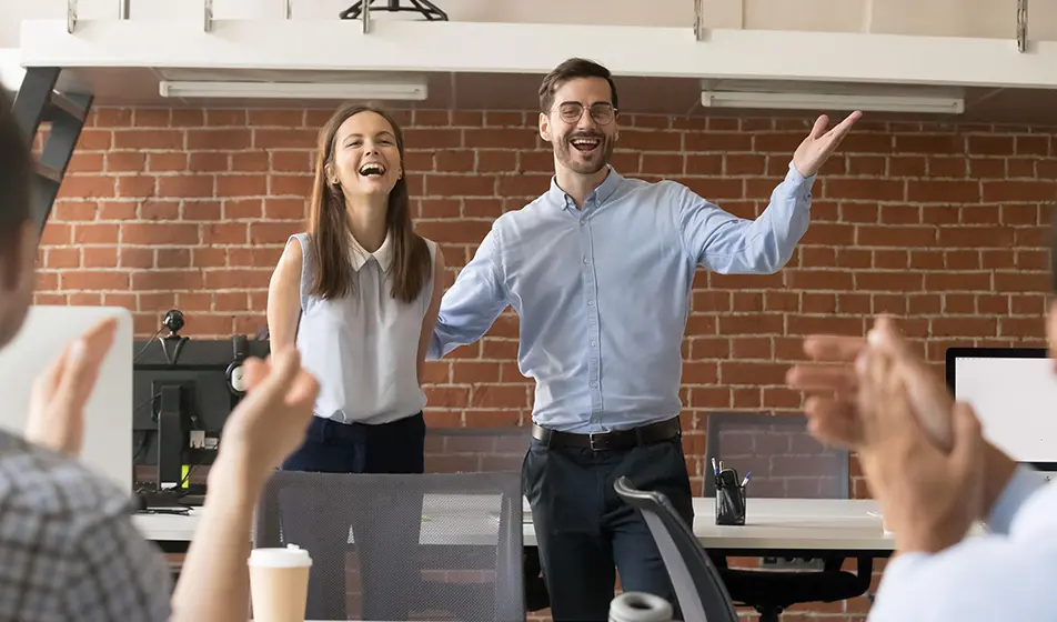 A new employee is being introduced at work. She is standing up in a modern office in front of her colleagues with her manager cheering and welcoming her to the team.