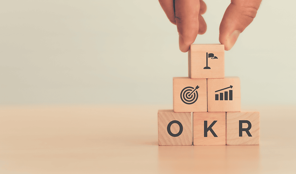 Small wooden blocks are built up into a pyramid shape. At the top of the pyramid is a cube with a flag symbol on. Underneath that block are two other blocks; one has a target symbol on it and the other has a growing bar chart on it. At the bottom of the pyramid are three blocks which spell out "OKR" which stands for "objectives and key results".