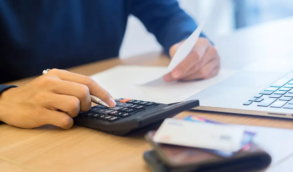 A man is using a black calculator to do payroll for his company. He is holding a slip of paper in one hand and his laptop is in front of him.