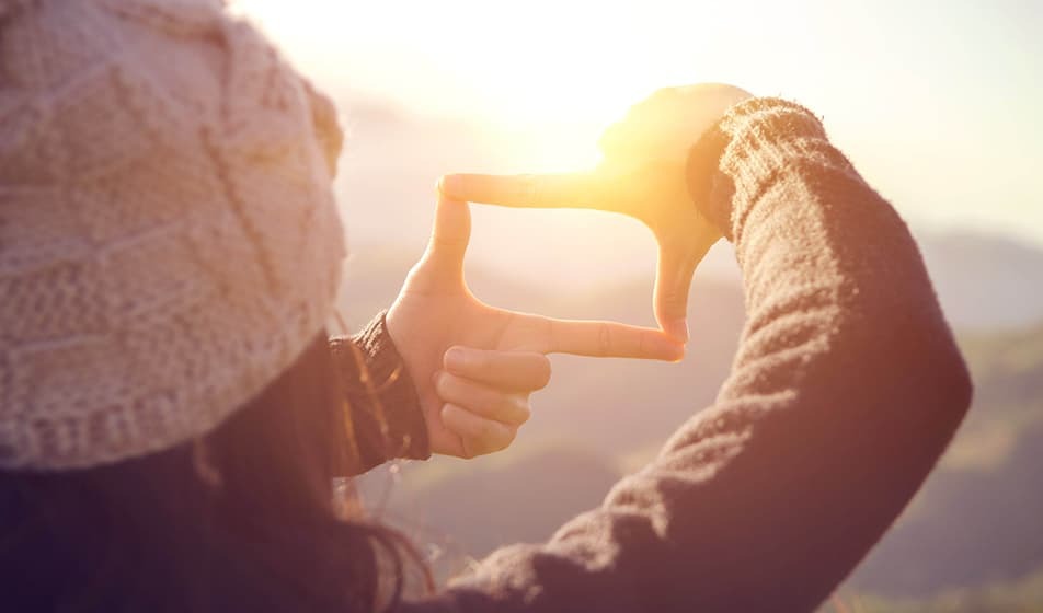 A woman in a fluffy hat is joining her two thumbs and index fingers together to create a rectangle shape. She is pretending to take a photo of the sunset.