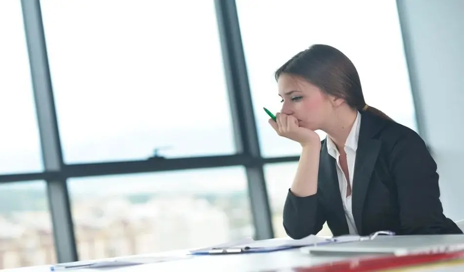 A business woman is sat at her desk surrounded by folders and paperwork. She is looking sad and tired with her hand covering her mouth.