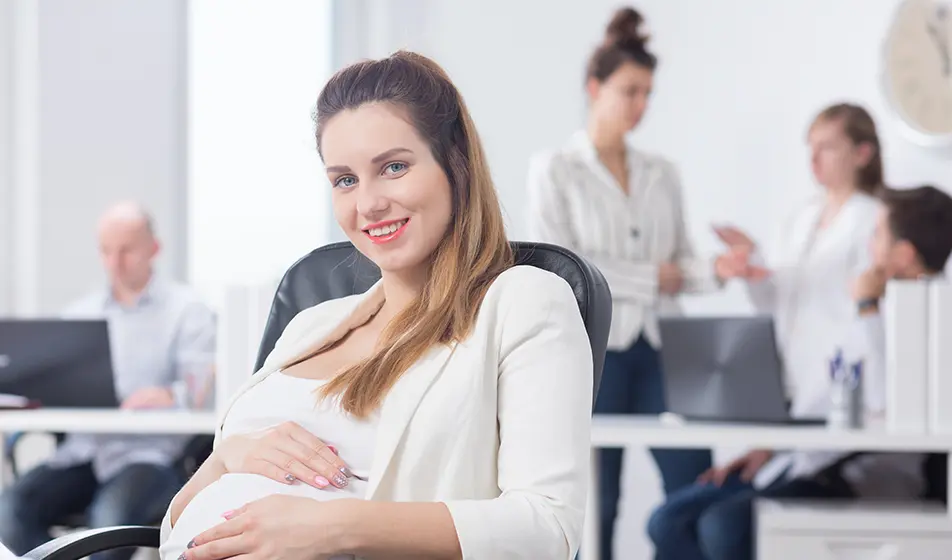 A business woman, who is pregnant, is smiling at the camera whilst holding her baby bump. She is sat in an office with her colleagues working behind her.