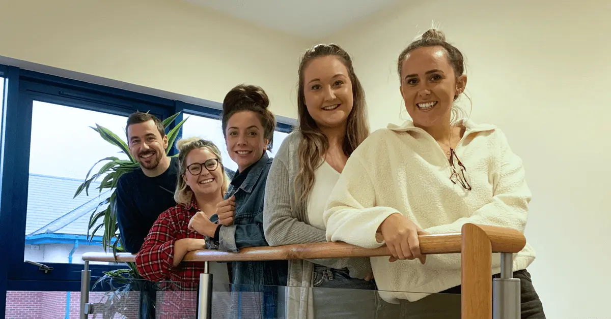 A self-managed team of five stand together at the top of a staircase holding the bannister and smiling at the camera.