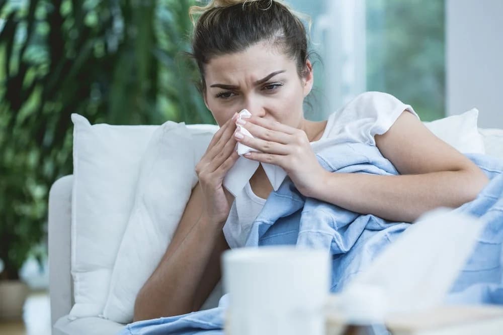 A woman is unwell and has a runny nose. She is lying down on a white sofa with a  blue blanket covering her. She is holding tissue to her nose.