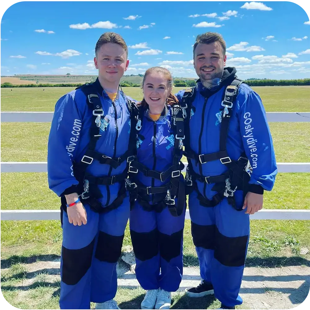 Two boys and one girl wearing blue all in one suits and harnessed up standing in a field ready to do their skydive