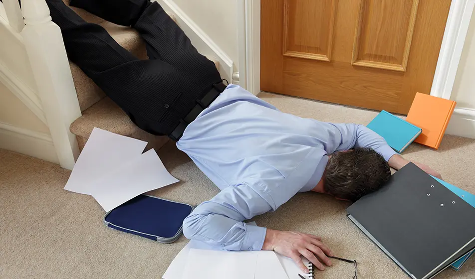 A businessman has fallen down the stairs and is lying face down into the carpet. He is surrounded by folders, books and his glasses which have fallen down the stairs with him.