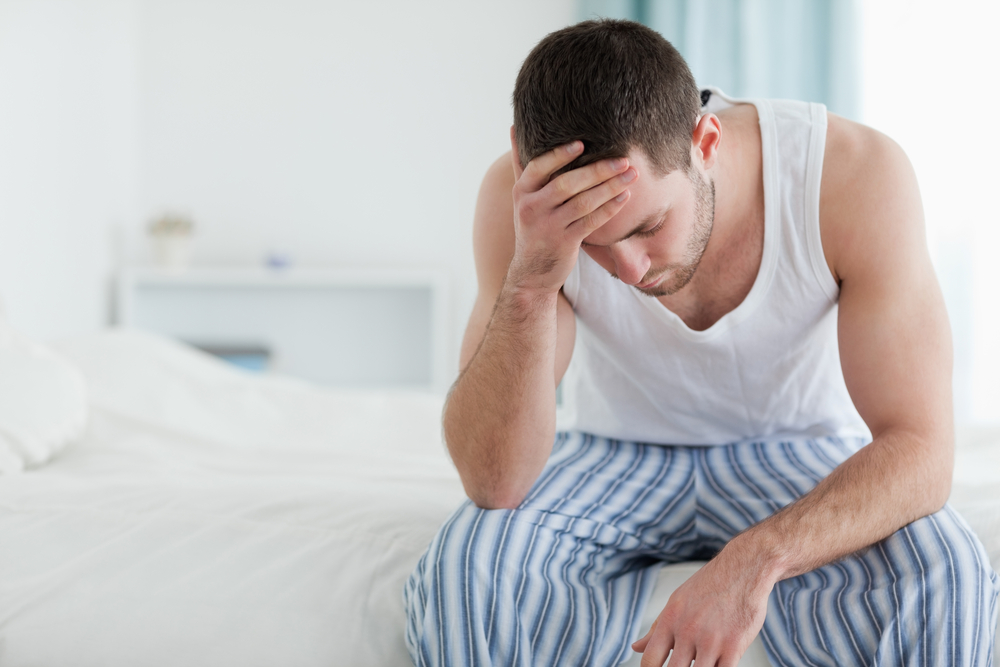 A man is wearing striped pyjama bottoms and a white vest top. He is sat on the edge of his bed with his hand on his head. He is feeling very unwell.
