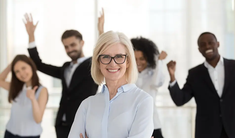 A business woman is wearing a blue shirt and is smiling at the camera. Behind her is blurry but four of her colleagues are stood cheering.