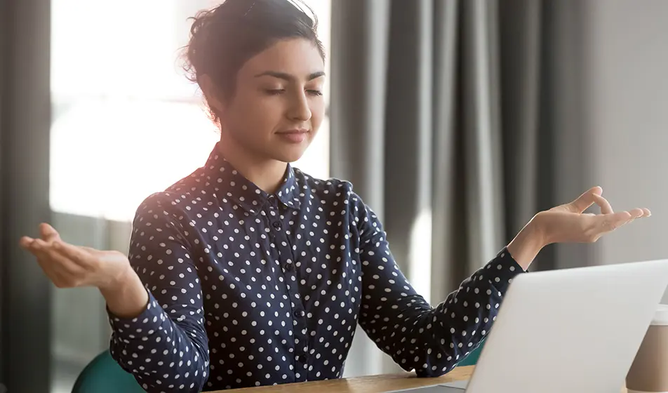 A woman is wearing a blue blouse with white polka dots. She has a laptop in front of her and is meditating.