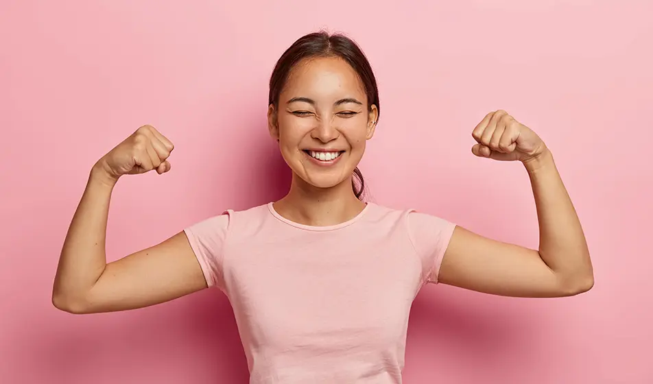 A woman in a pink shirt is stood against a pink wall. She is flexing her arms and smiling at the camera with her eyes closed.