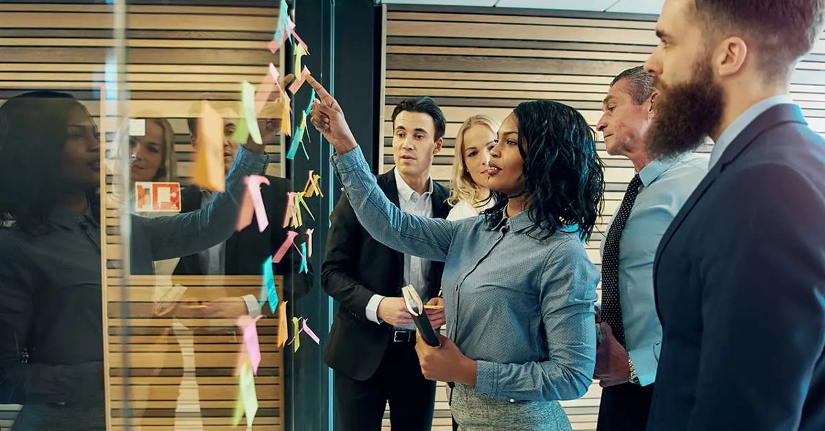 A team of five colleagues are organising a work schedule on a glass wall using sticky notes.