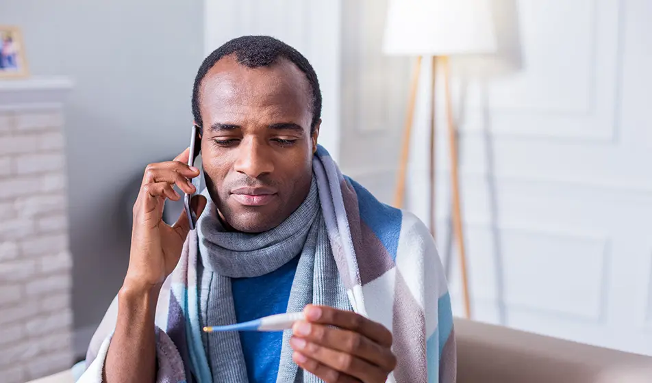 A man is feeling unwell and has taken his temperature. He is wearing a scarf and a blanket whilst calling his work to tell them he is sick.