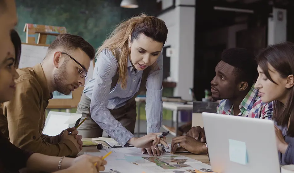 Six colleagues are gathered around a wooden table together. They have a laptop on the table as well as paperwork showing architectural plans.