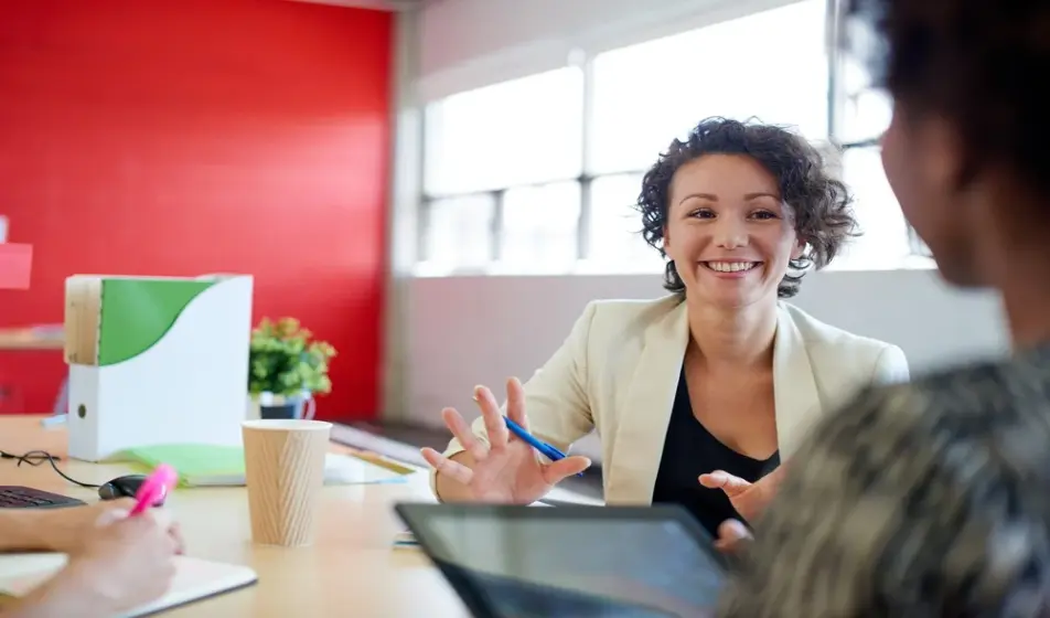 A happy woman is smiling in a meeting with her colleague who is holding an iPad. There is a bright red wall behind her.