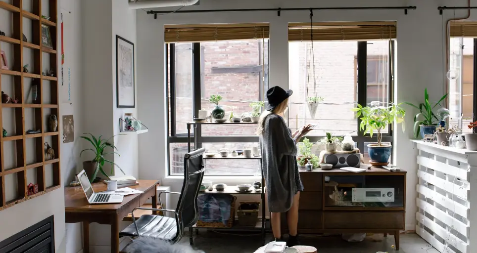 Woman with hat standing in fron of a window using a tablet device surround by modern room with house plants and workstation