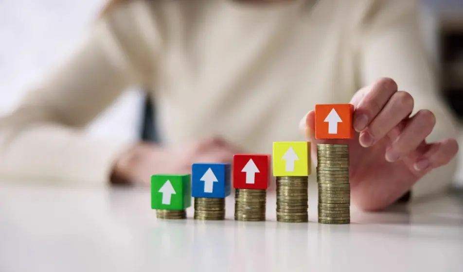 A hand placing a block with an upward arrow on a stack of coins, illustrating increasing employer National Insurance costs