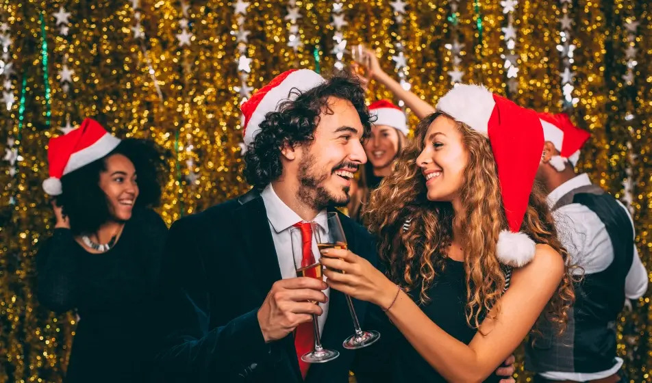 People dancing with Santa hats on in the background, a man and a woman in the front of the picture, hugging and cheering with Champagne 