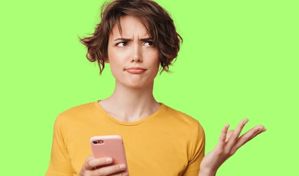 Confused young woman in a yellow shirt holding a smartphone, raising her hand in uncertainty against a bright green background.